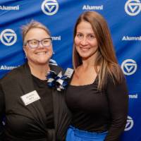 Two colleagues smile in front of the GVSU Alumni backdrop